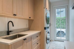 Laundry room built ins in white oak with a stone counter, sink and stacked washer dryer tower