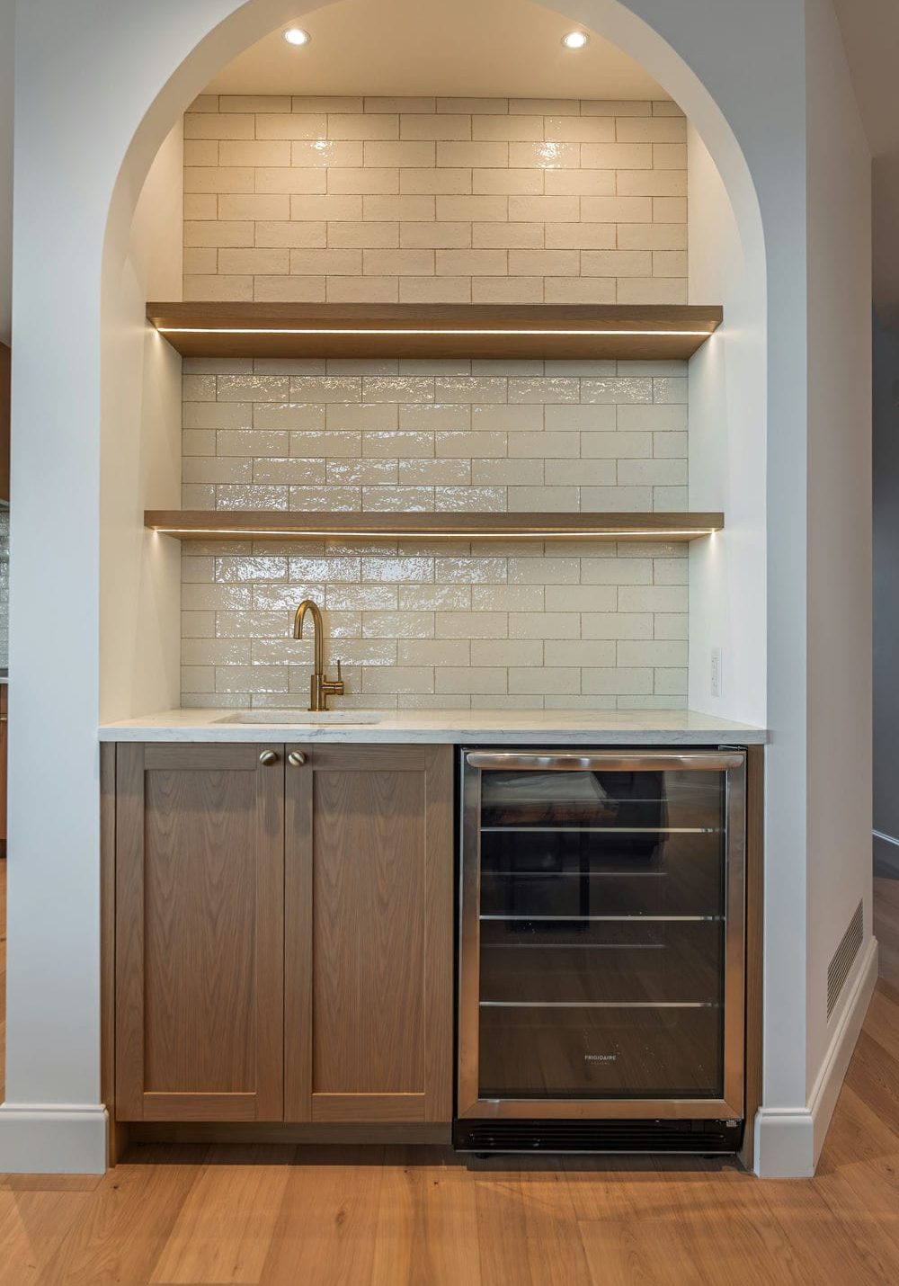 Arched wet bar niche with stained oak cabinets, lit floating shelves and a built in wine fridge